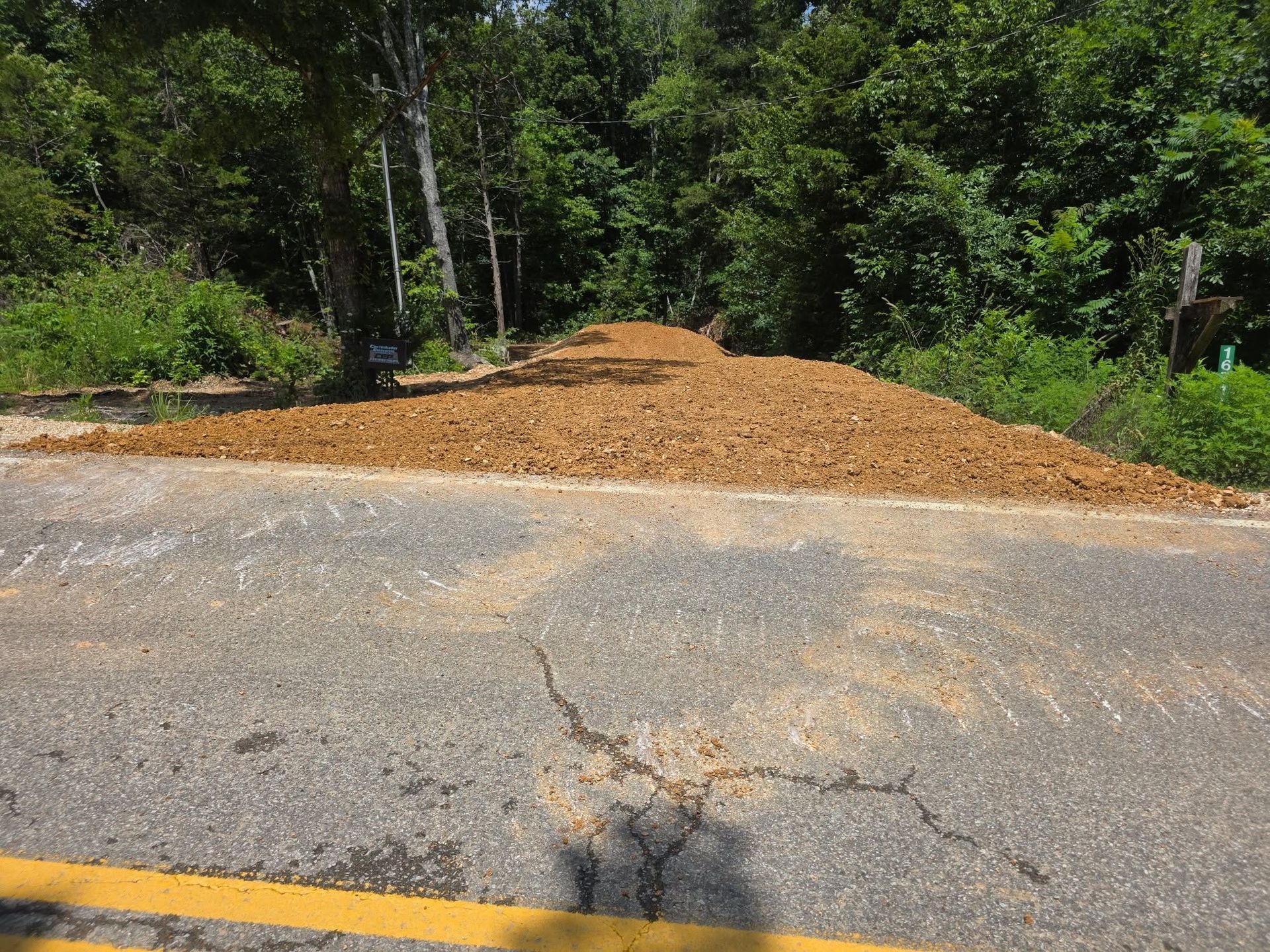 Piled tan gravel beside a cracked roadside in front of dense green trees
