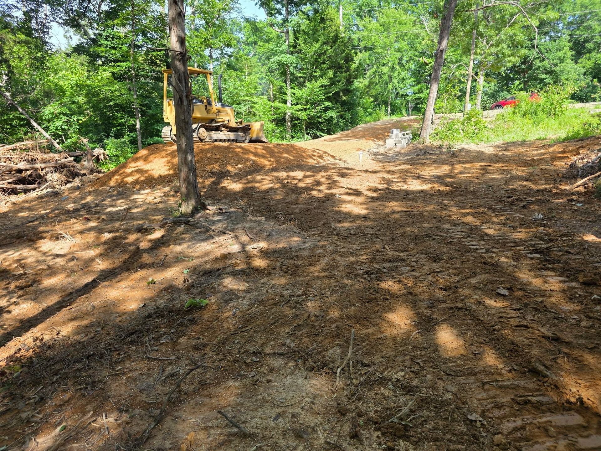 Cleared dirt path through a wooded area with a yellow excavator in the background