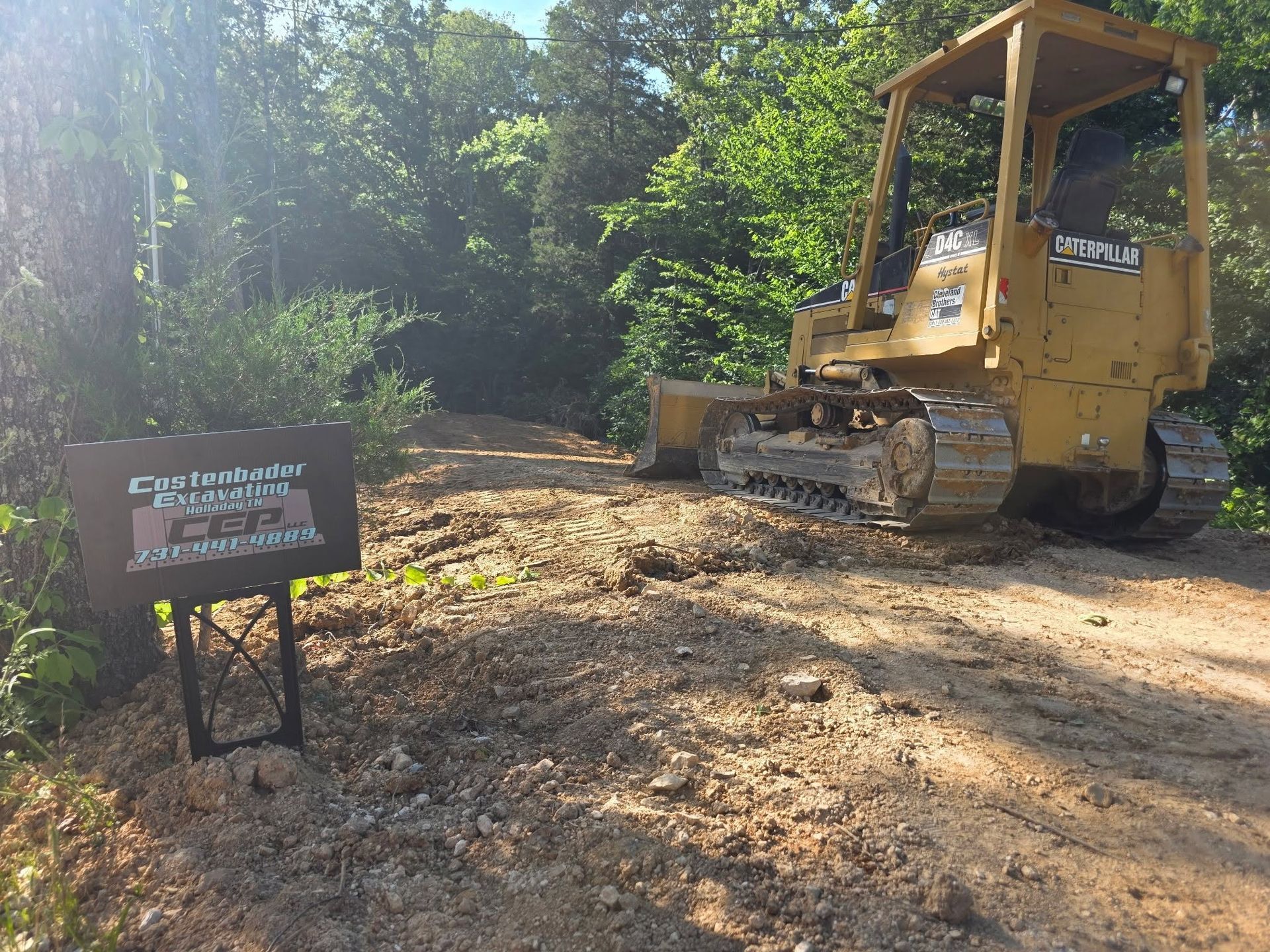 Yellow bulldozer clearing a wooded dirt lot beside a warning sign.
