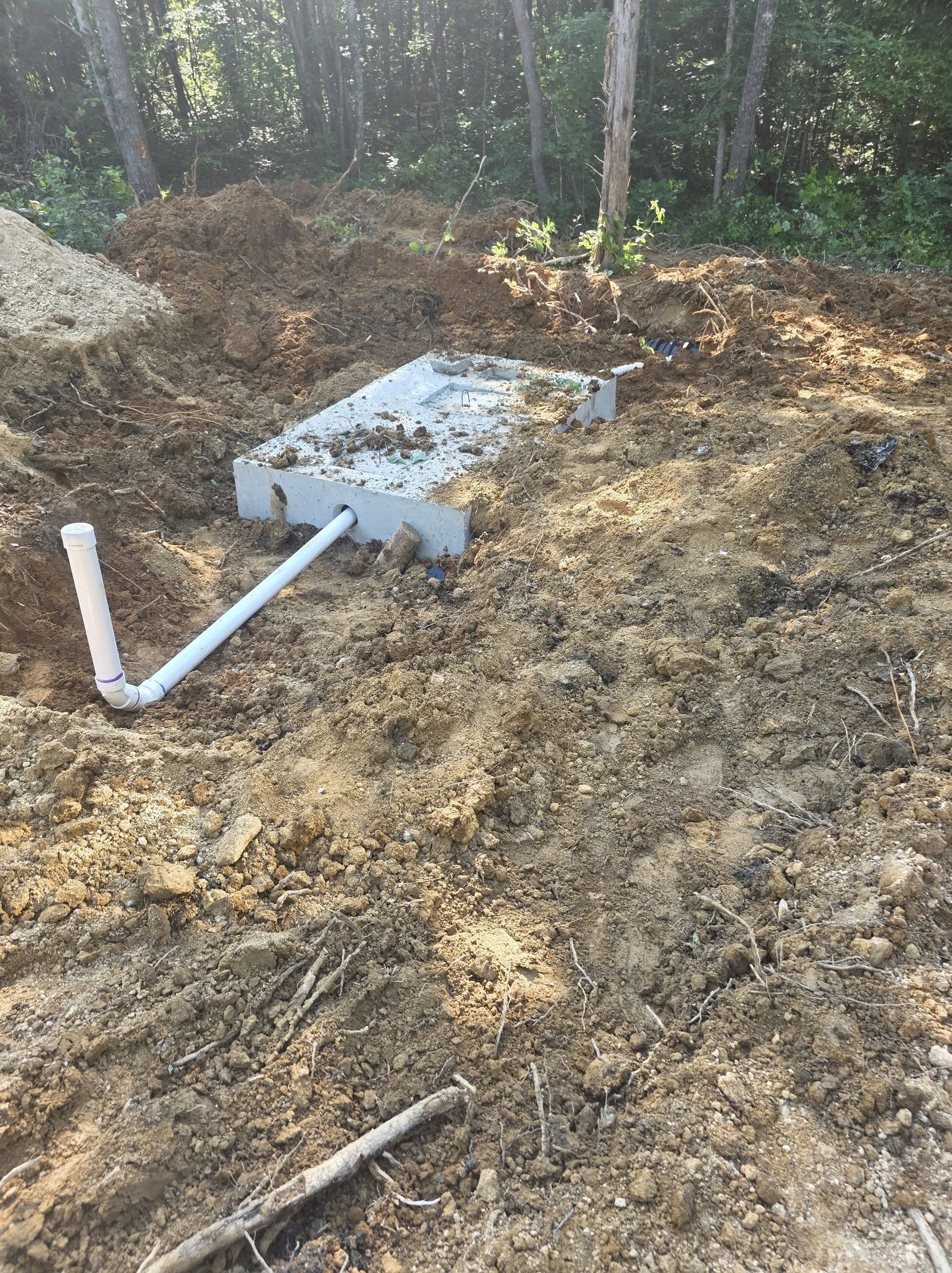 Installed septic tank and white PVC pipe in a dirt excavation near trees