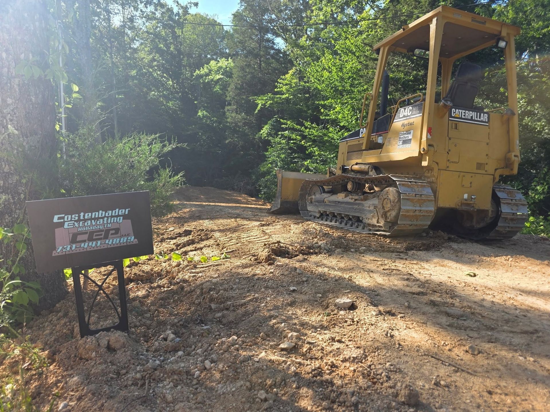 Yellow bulldozer on a dirt clearing beside a sign, surrounded by trees and sunlight