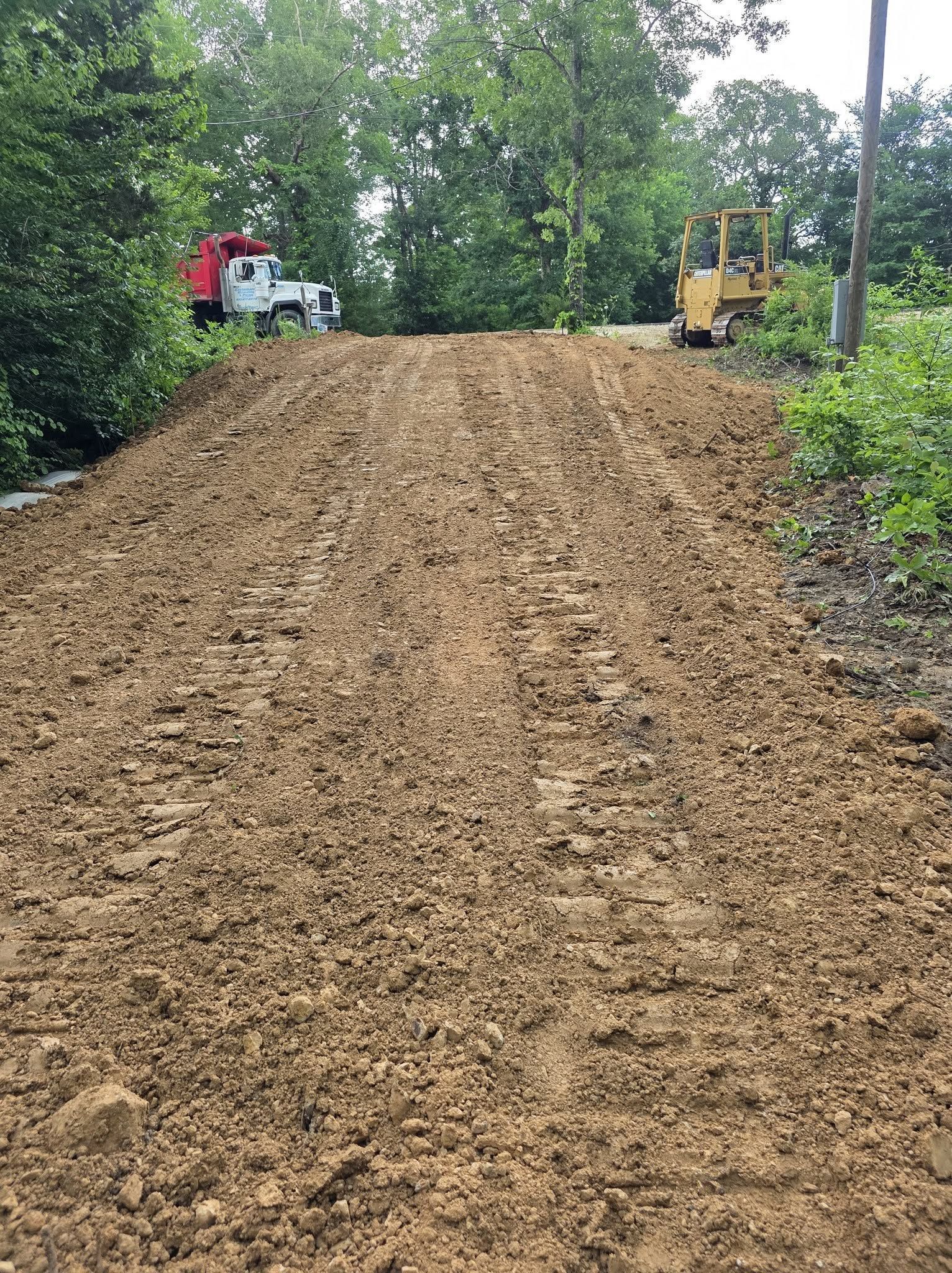 Freshly graded dirt road on a wooded hill with a red truck and yellow bulldozer at the top