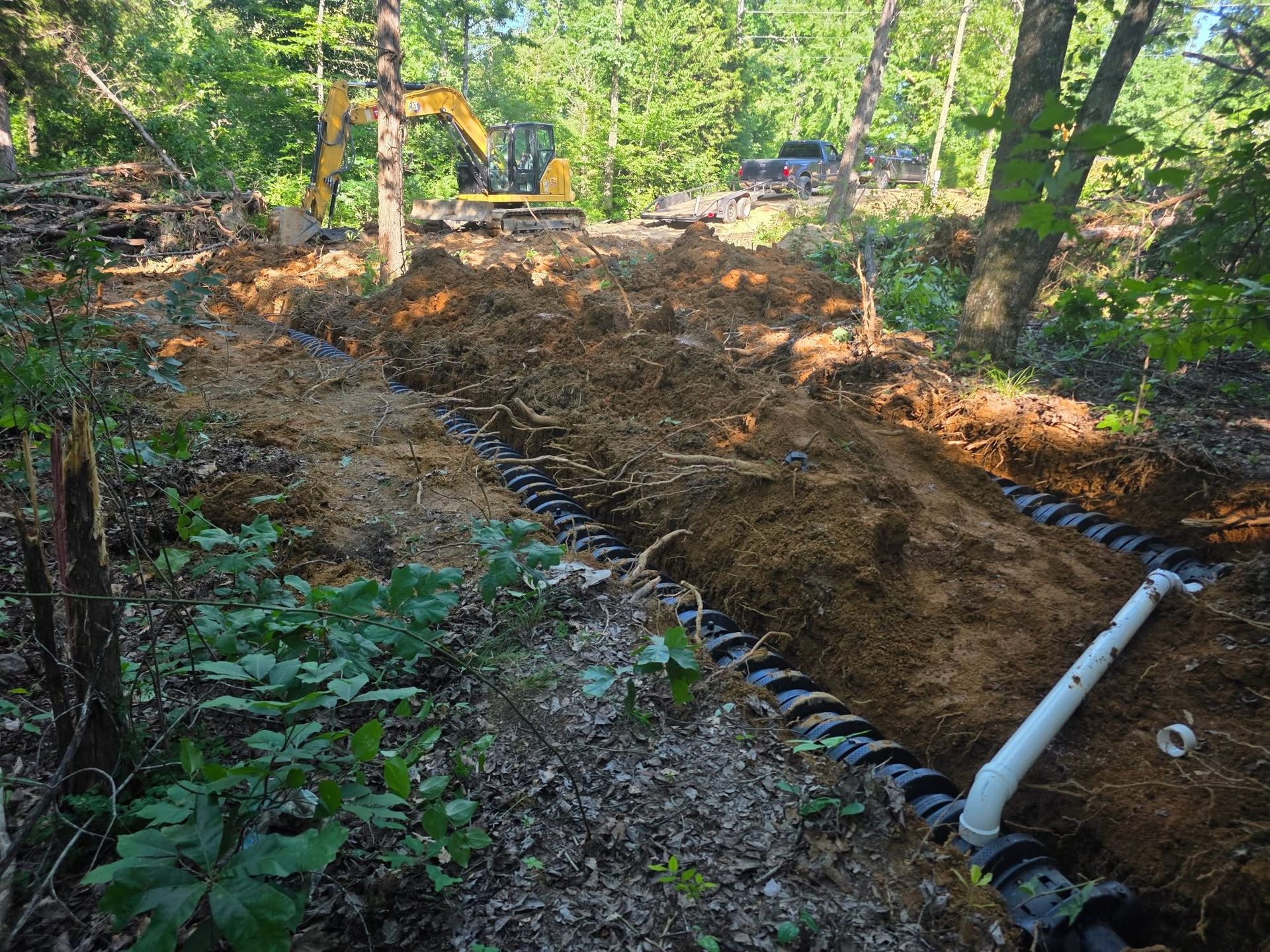 Excavator digging a trench in a wooded area, with white drainage pipe along the ground.