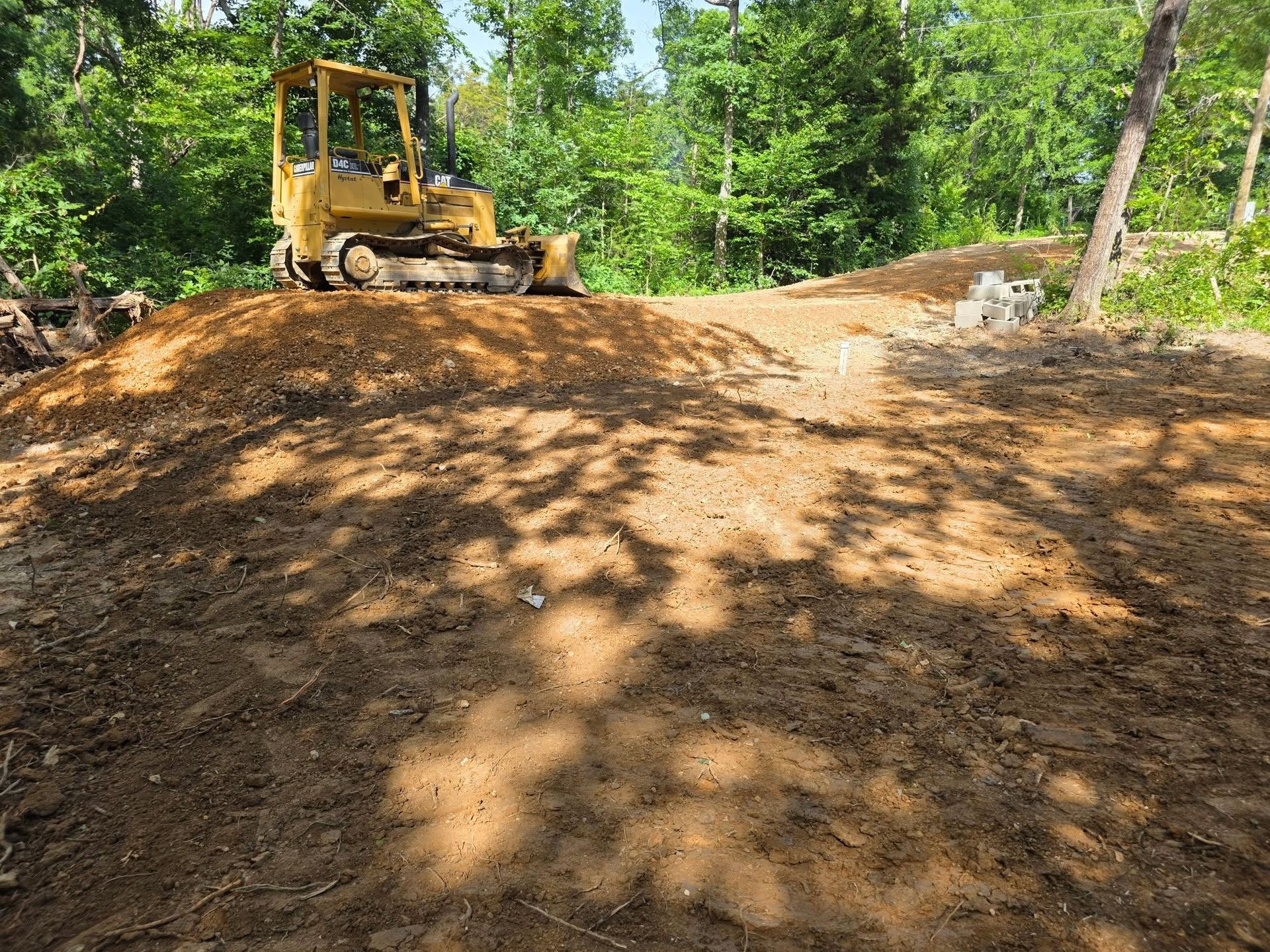 Yellow bulldozer on a dirt mound in a wooded clearing under bright sunlight
