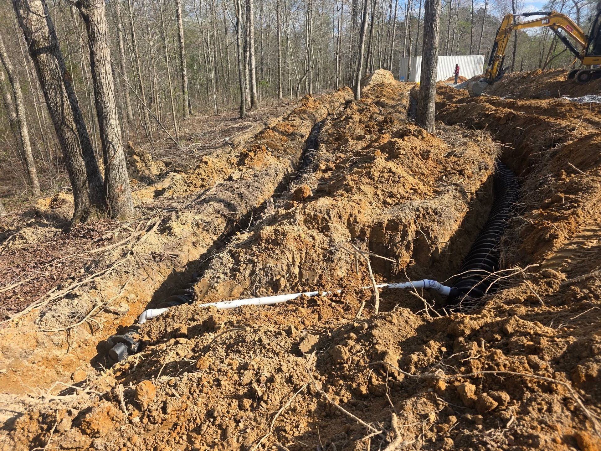 Excavator digging a deep trench in a wooded area with piles of fresh earth.