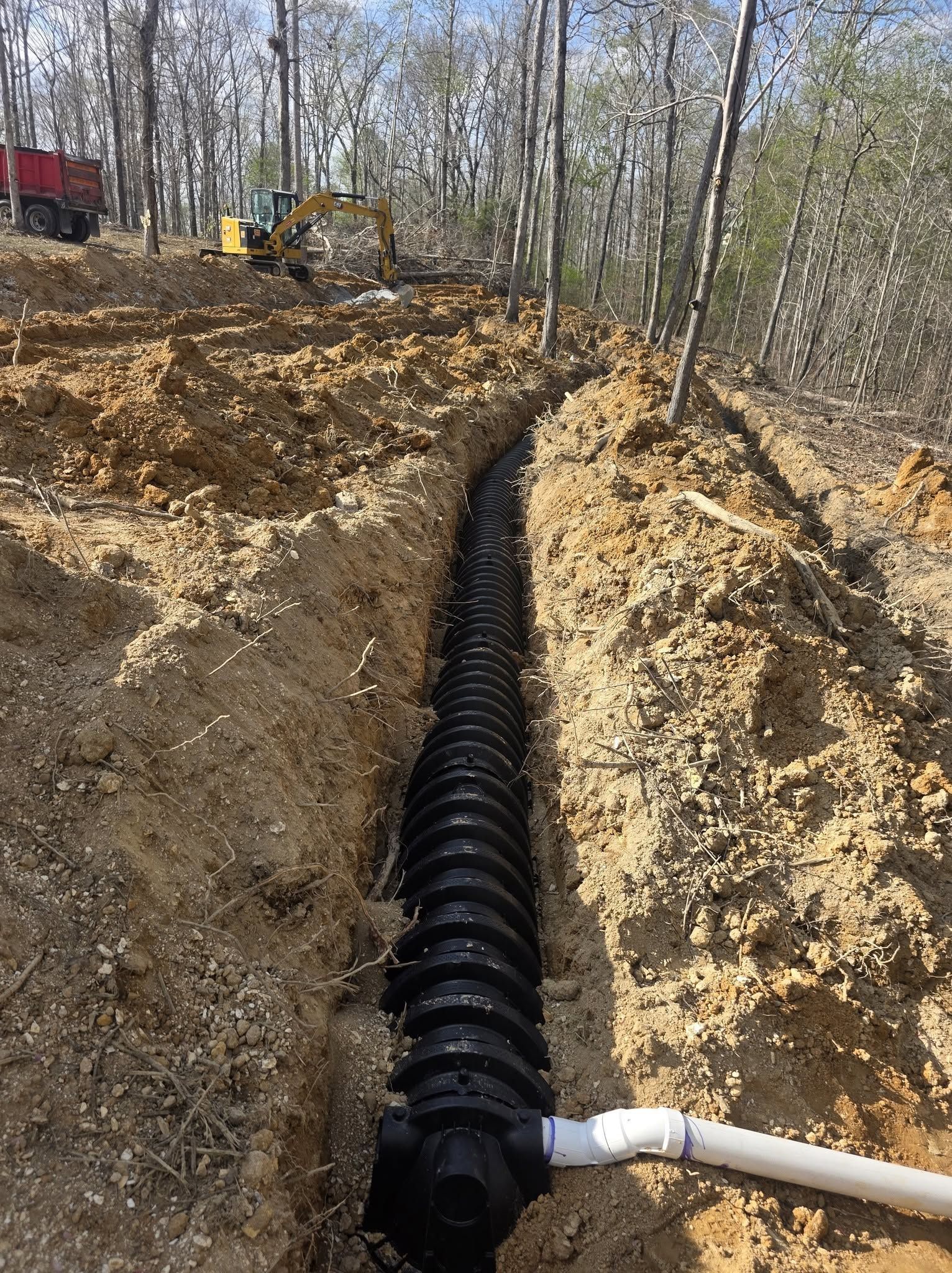 Construction trench with black drainage pipe and white elbow pipe in a wooded, dirt excavation area