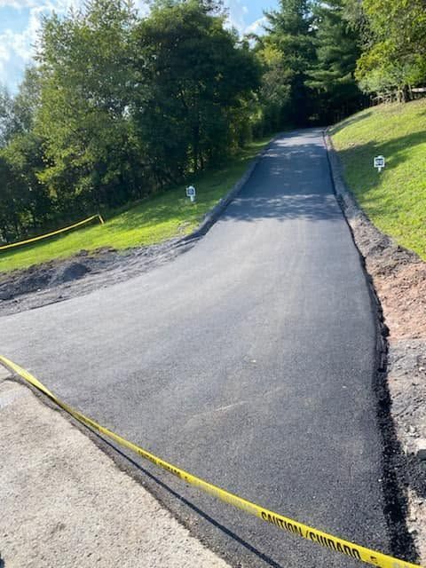 Freshly paved driveway or road with yellow caution tape, bordered by grass and trees on a sunny day