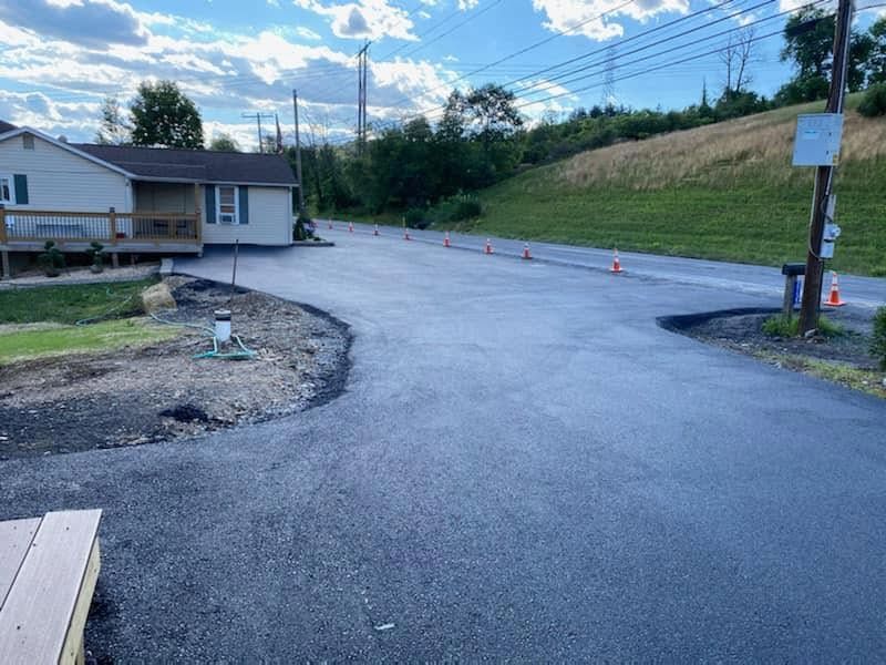 Freshly paved curved driveway beside a house, with cones, a grassy embankment, and a blue sky.