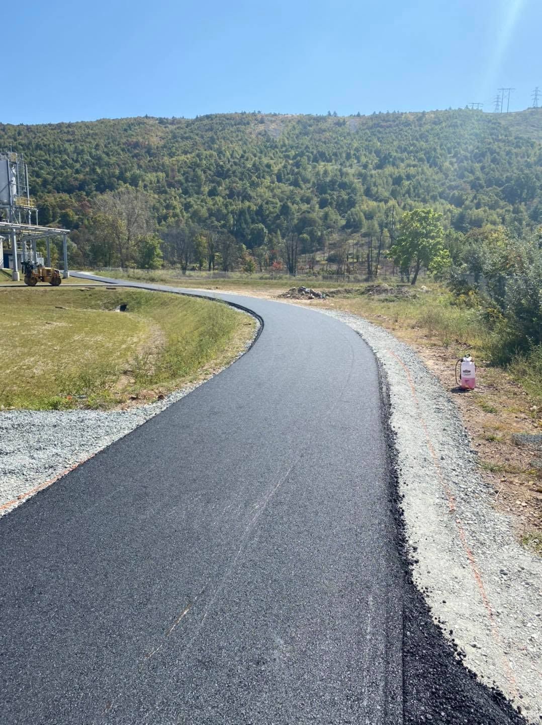 Curving black paved path beside gravel and grass, leading toward wooded hills under a bright sky