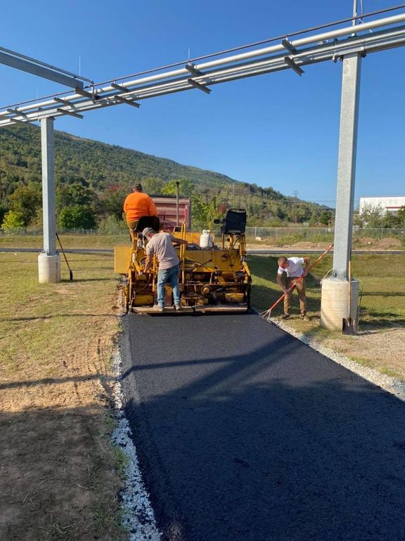 Workers paving a narrow road under a blue metal frame in a rural field