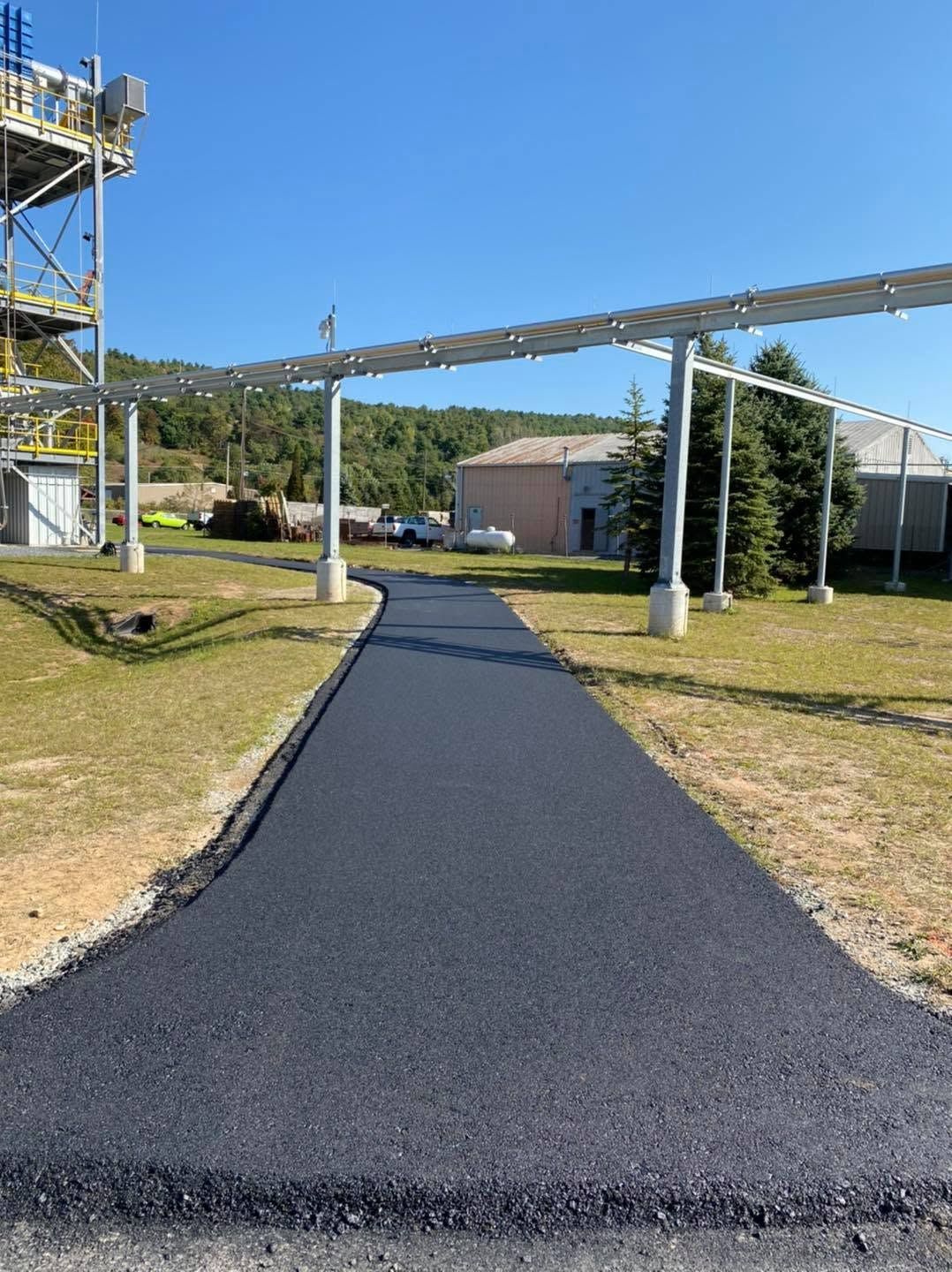 Freshly paved black pathway leading to a small building, with grass and metal structures on both sides.