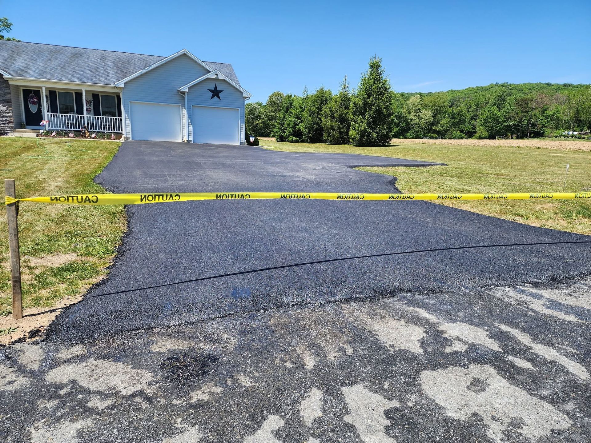Driveway and garage of a house blocked by yellow caution tape, with fresh black asphalt paving.