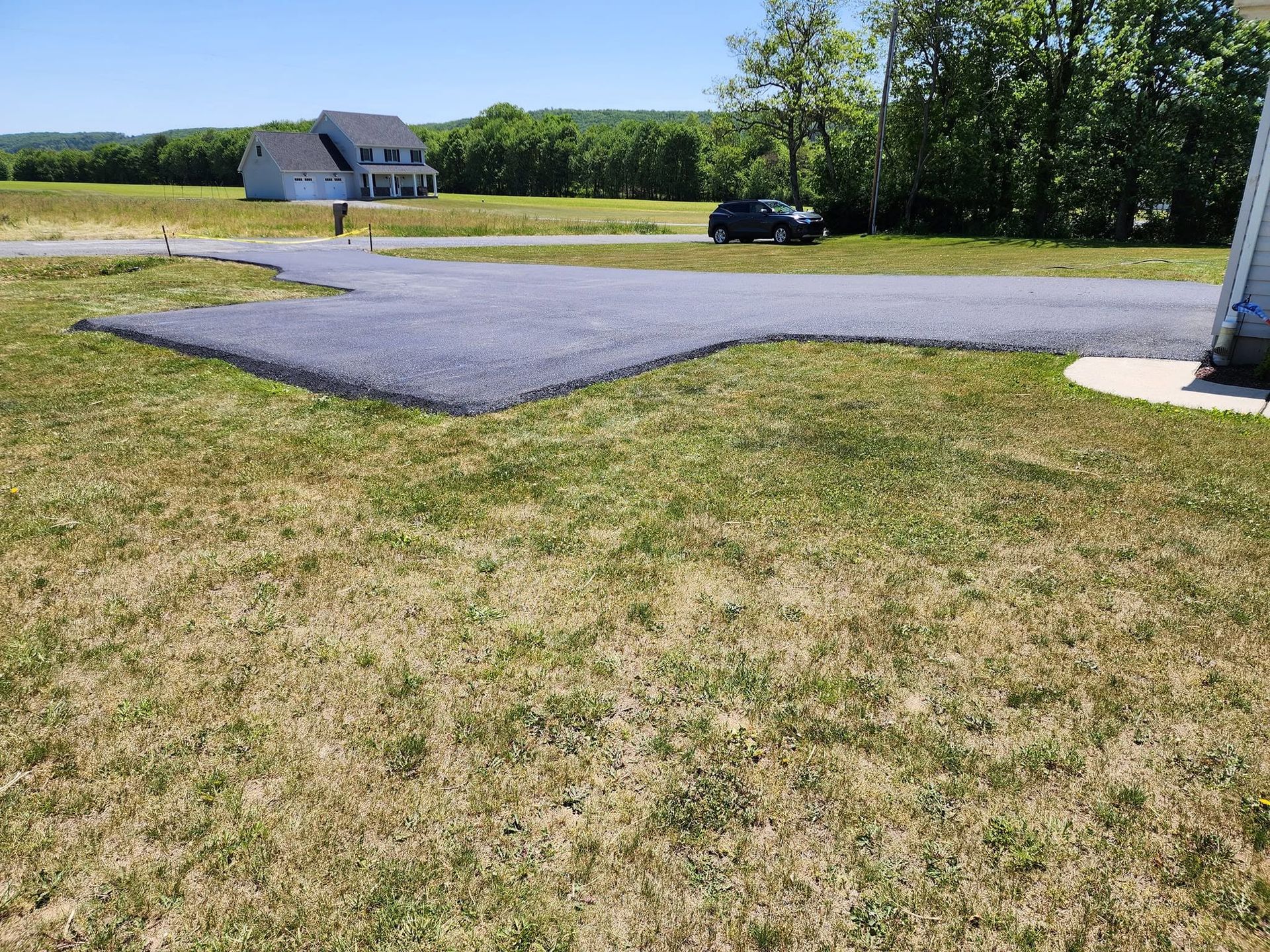 Grassy yard with a paved driveway, house in the distance, and a parked car near trees under a clear sky