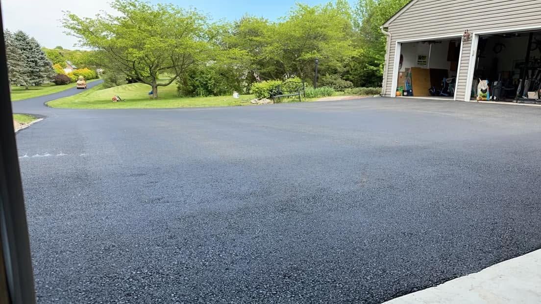 Freshly paved dark asphalt driveway beside a garage, with green trees and a sidewalk in the background