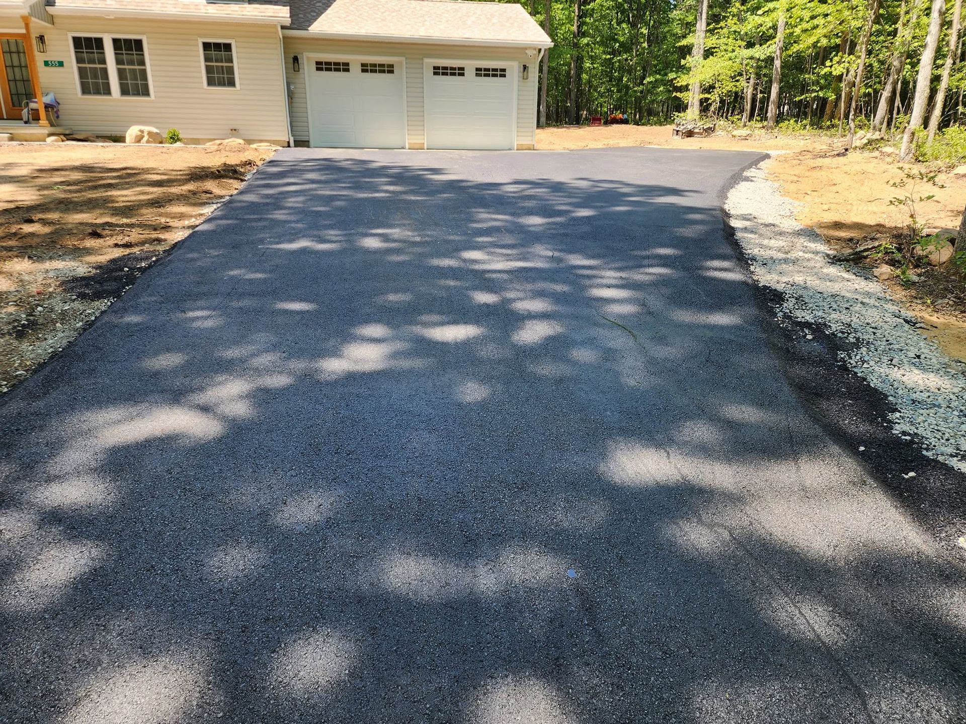 Freshly paved dark driveway leading to a beige house with a double garage, surrounded by trees