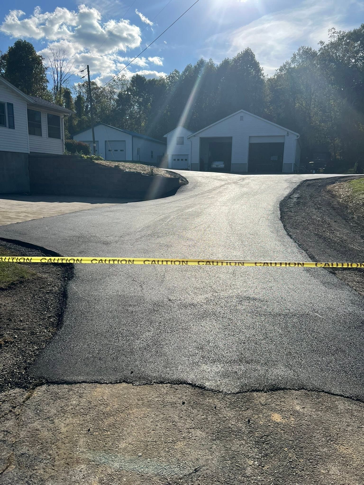 Freshly paved driveway rising to a garage, with yellow caution tape in front under bright sun.