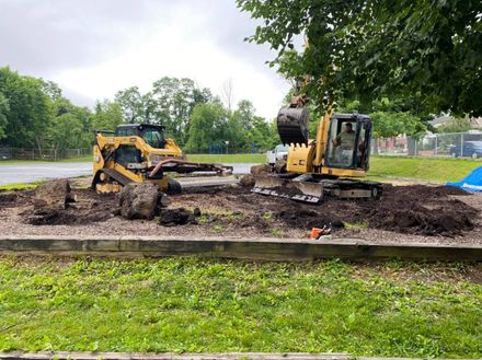 Two yellow excavators digging dirt beside a wet park path under trees