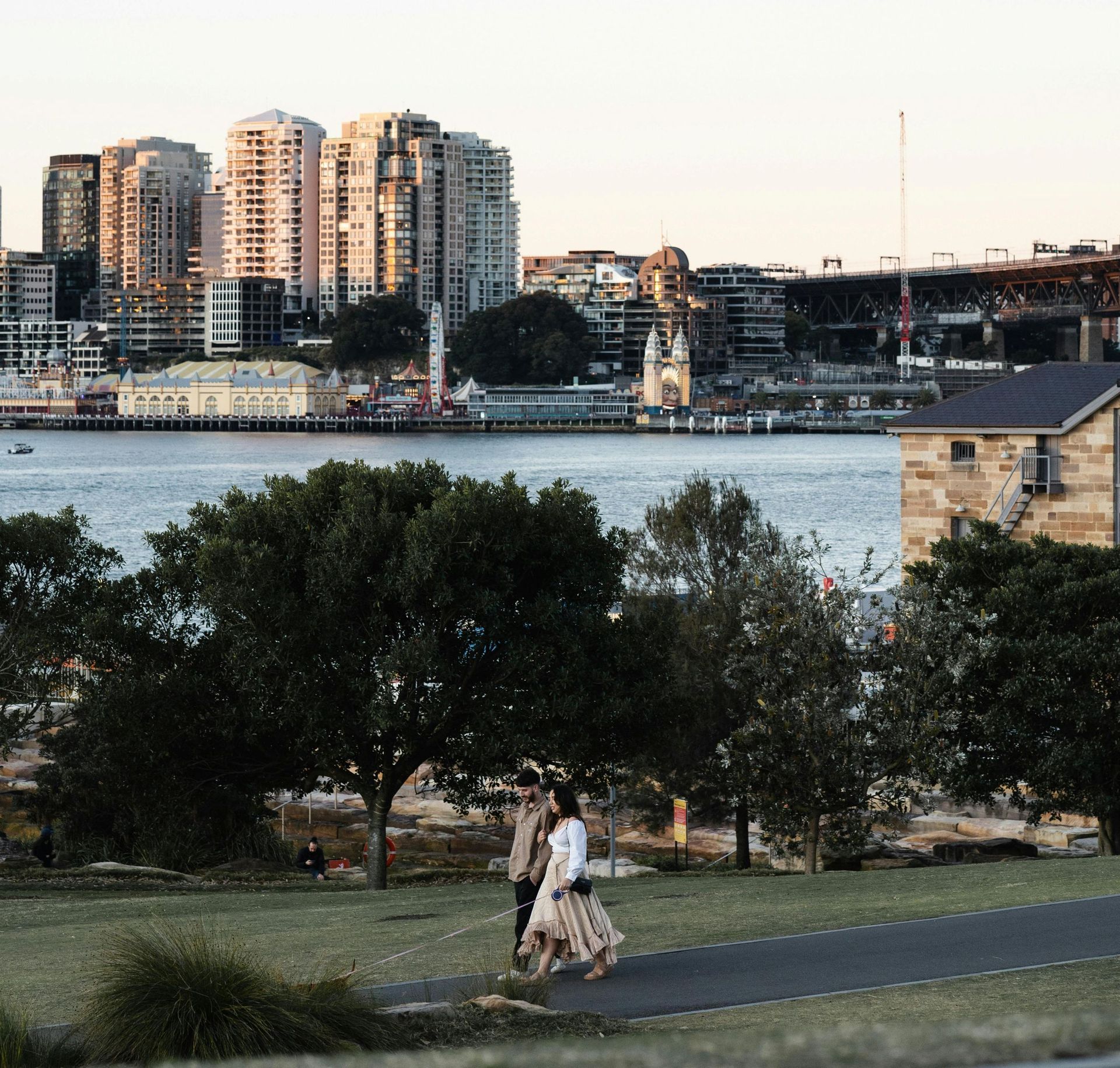 Two people walking on a grassy park path by a river, with city buildings and a waterfront skyline beyond.