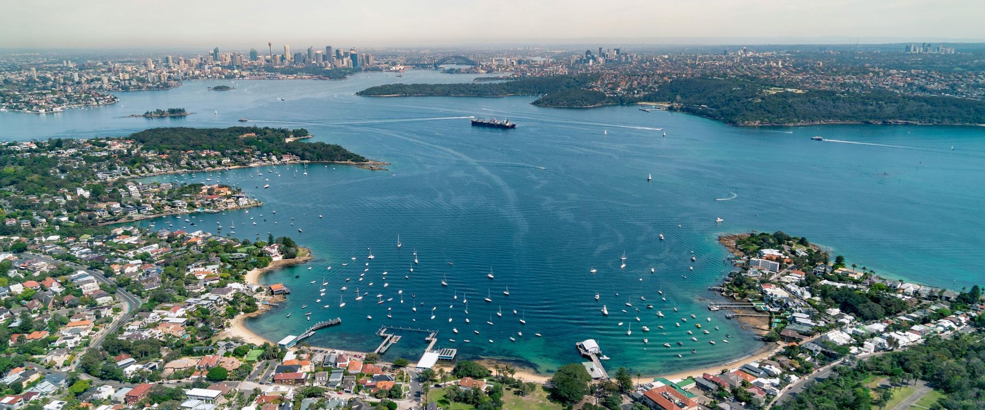 Aerial view of a blue bay with boats, surrounded by dense coastal neighborhoods and a distant city skyline