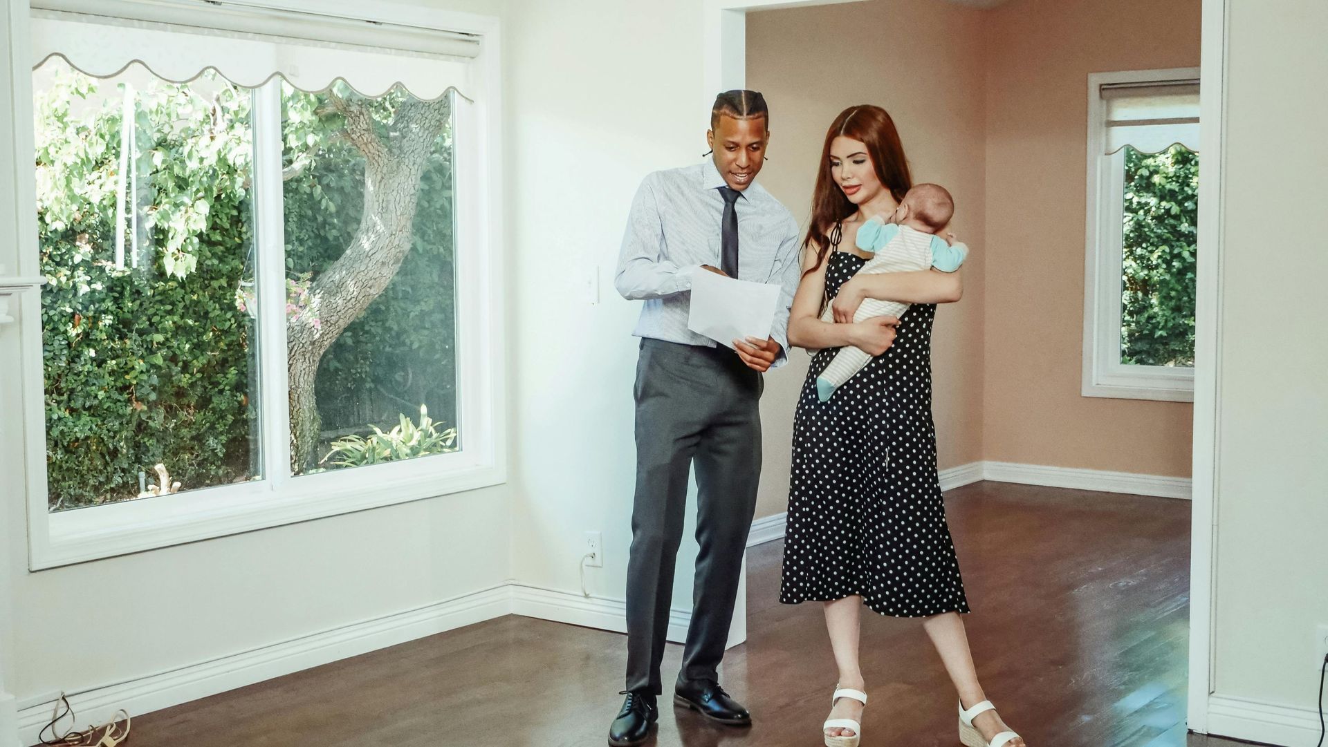 A real estate agent stands with a person holding a baby in an empty room with wood floors and large windows.