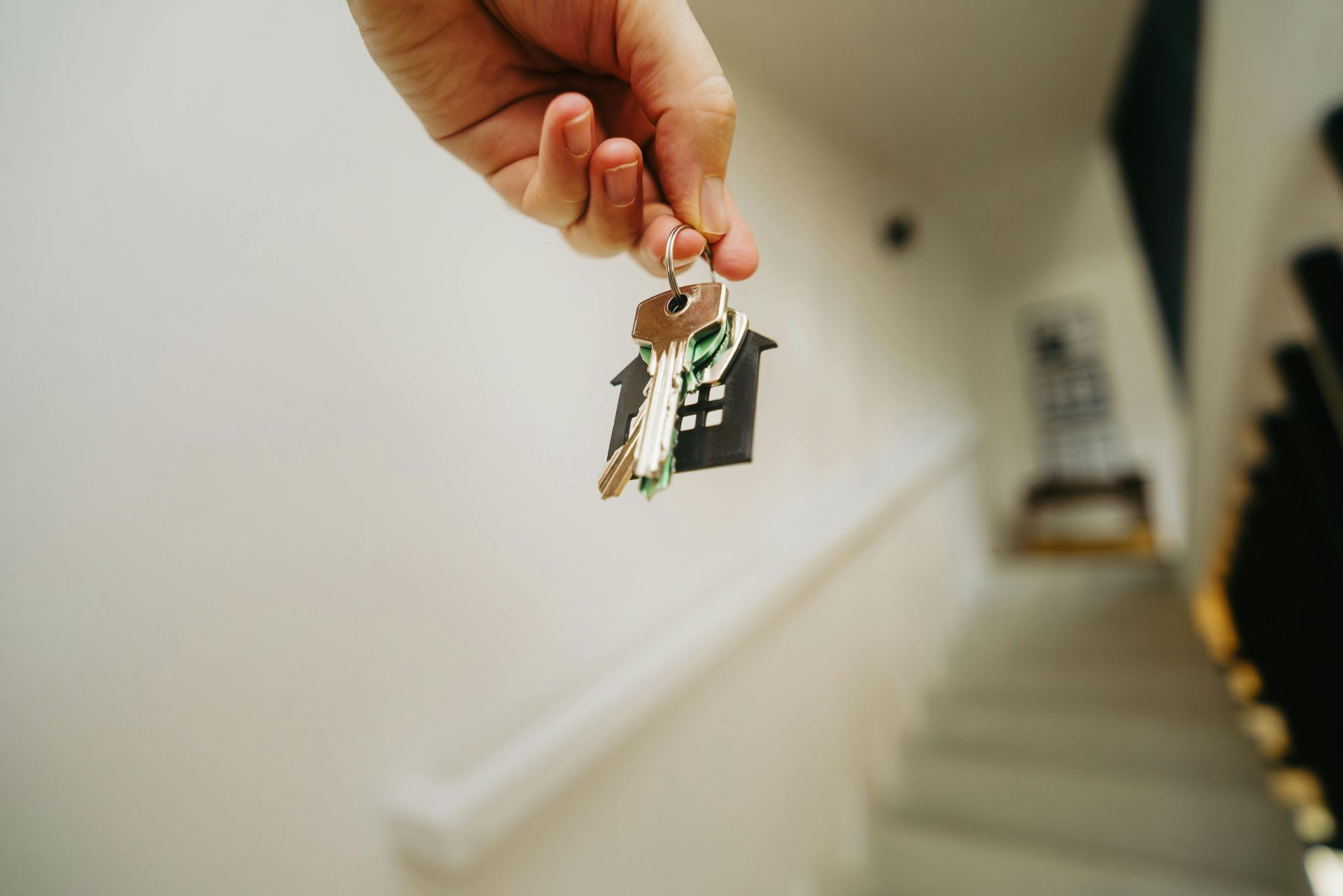 A hand holds up a set of keys with a house-shaped keychain against a blurred background of a white wall and staircase.