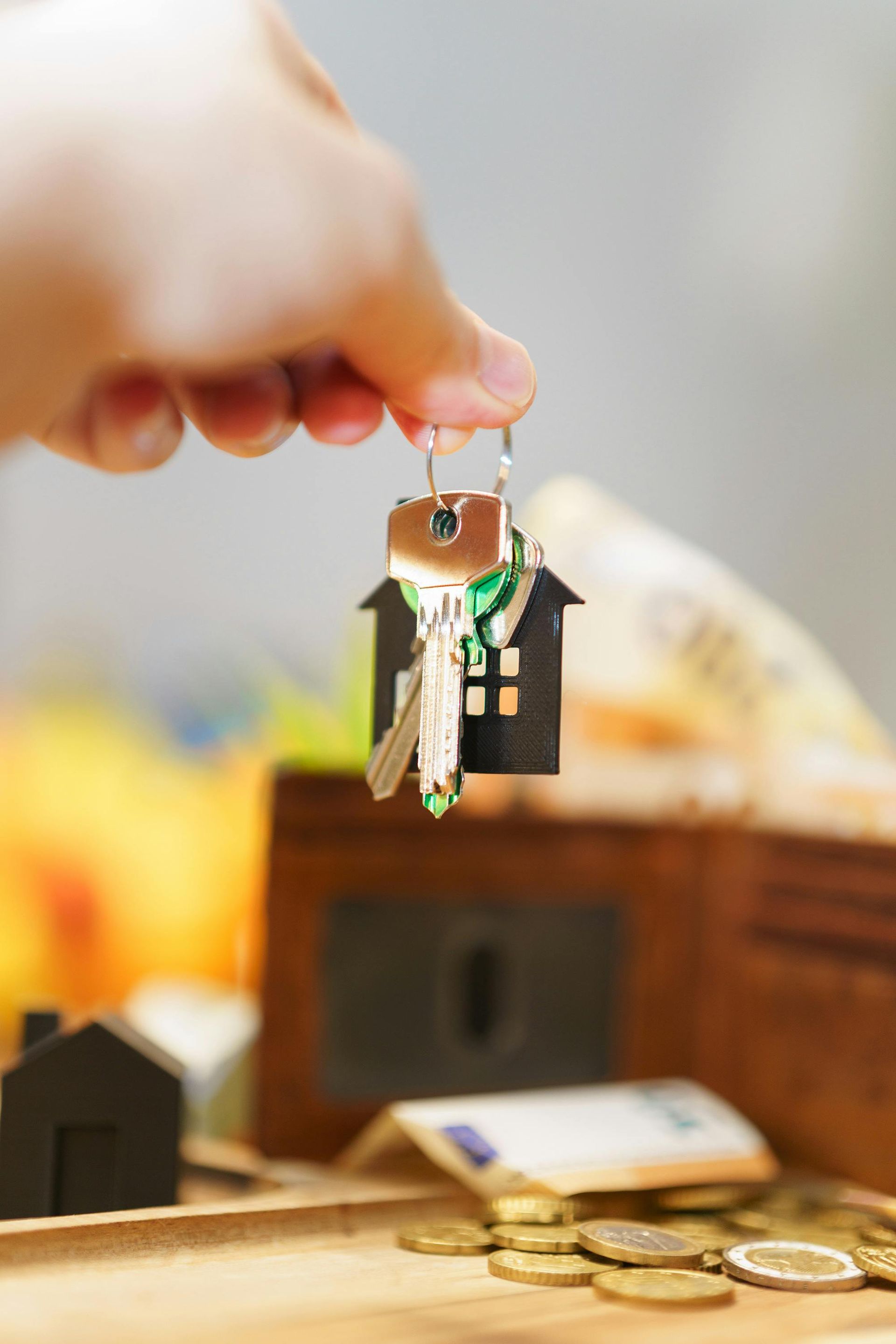 Hand holding a small keychain house with green roof above scattered coins on a table