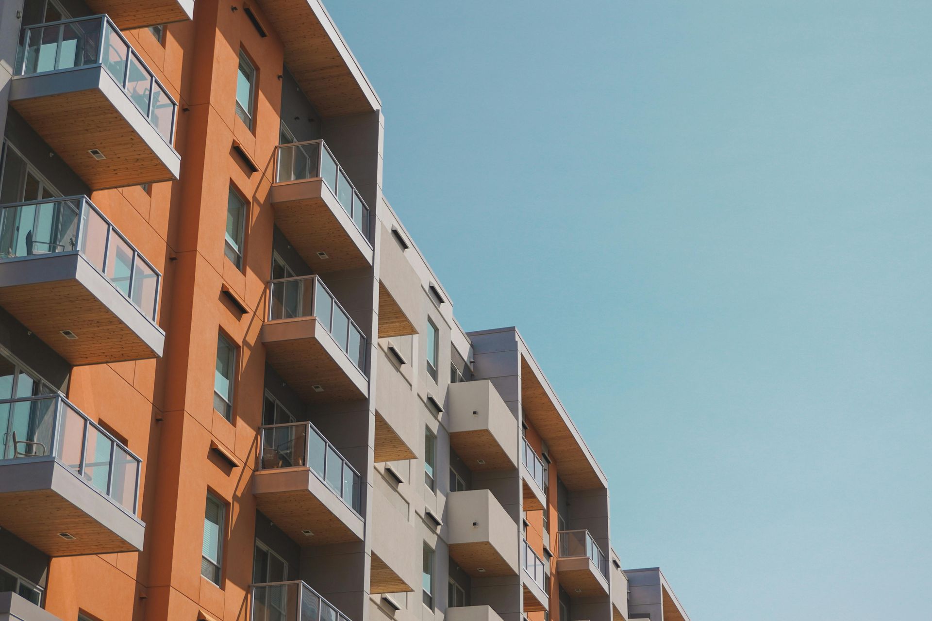 Low-angle view of a multi-story apartment building with balconies, orange and grey walls, against a clear blue sky.