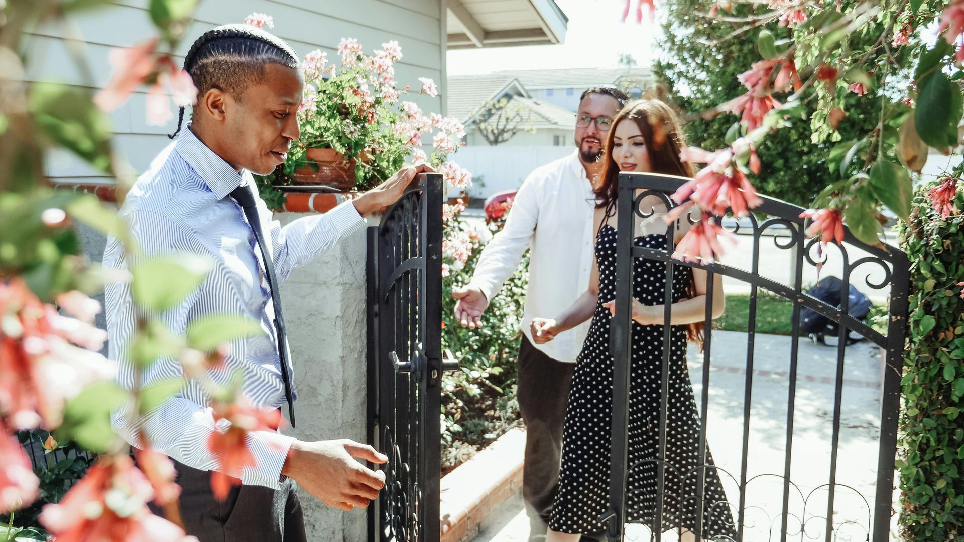 People standing by a garden gate at a sunny outdoor event, with flowers and a house in the background.