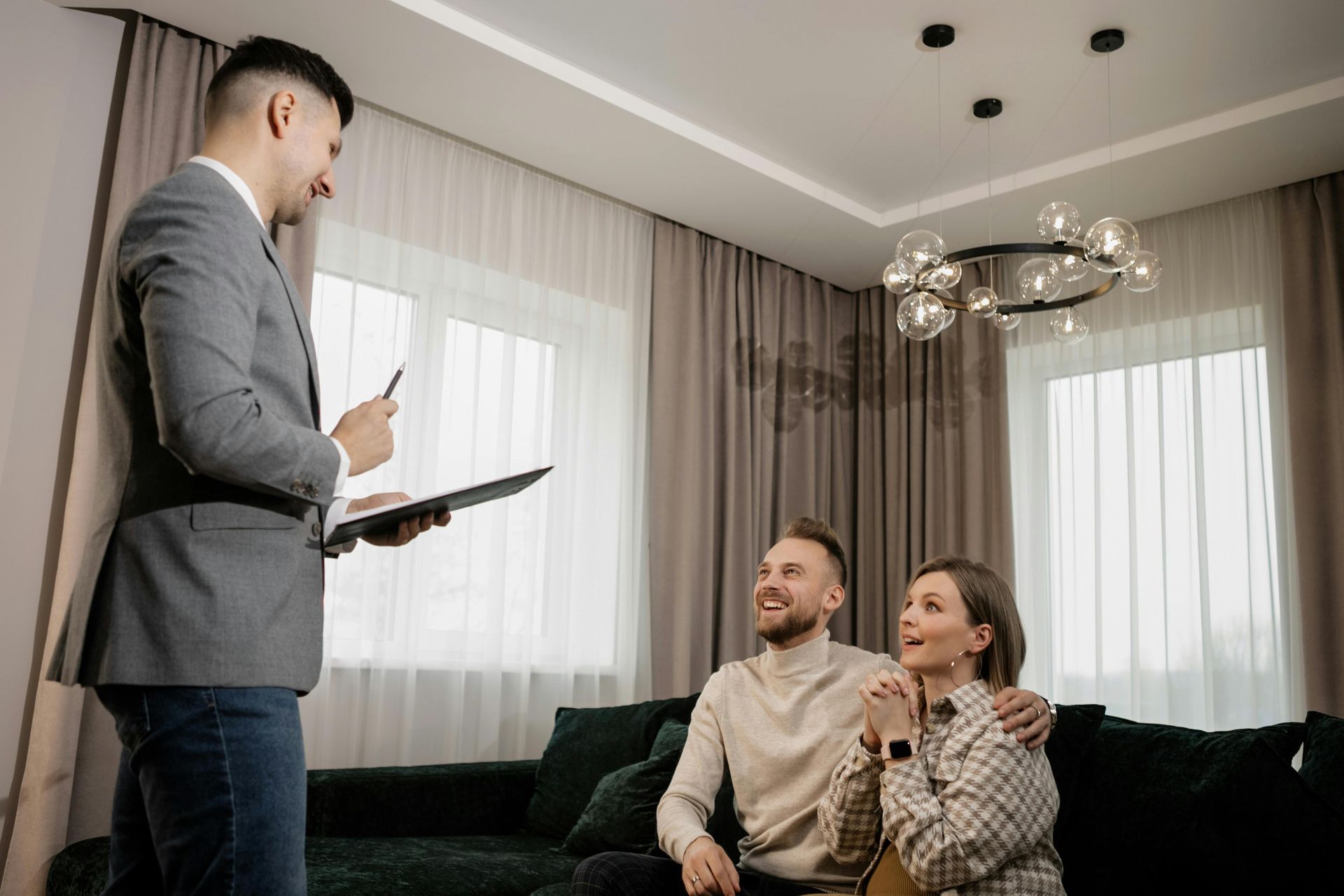 Real estate agent showing a couple a bright living room with large windows and a dark sofa