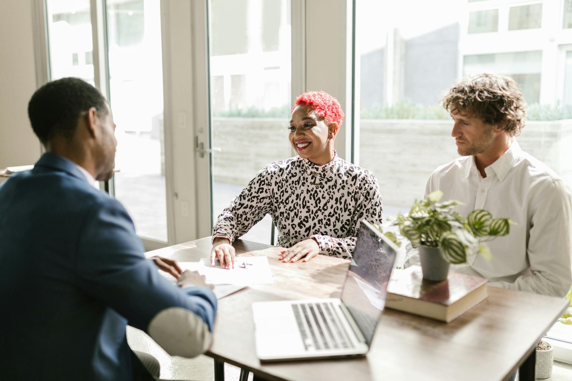 Three people in a bright office meeting, smiling across a table with laptop and papers.