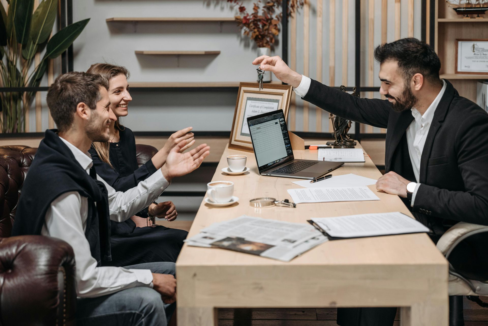Three coworkers in a meeting at a conference table, one holding up a tablet in a modern office.