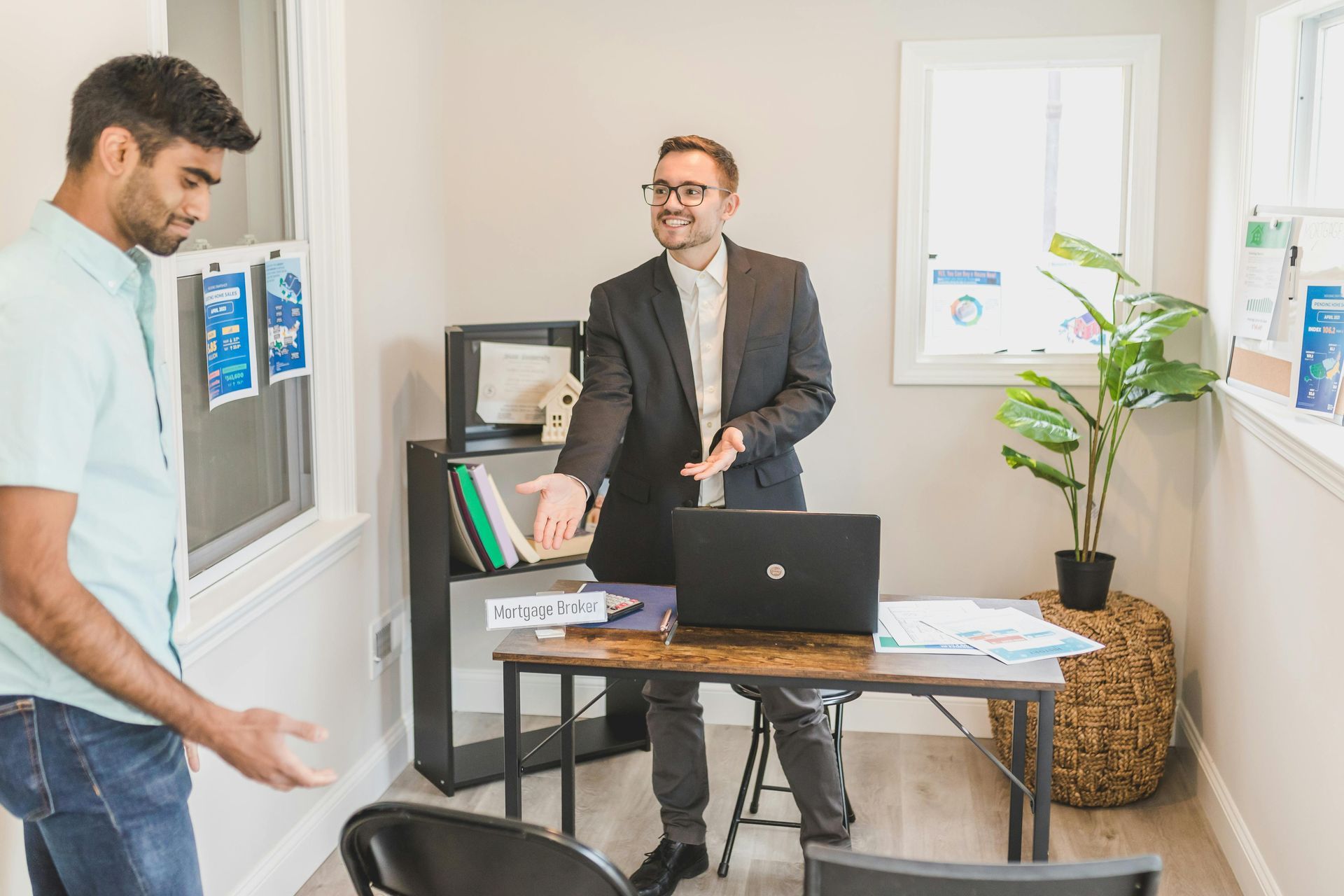 Two people in a bright office meeting, one standing by a laptop and desk with papers and plants.