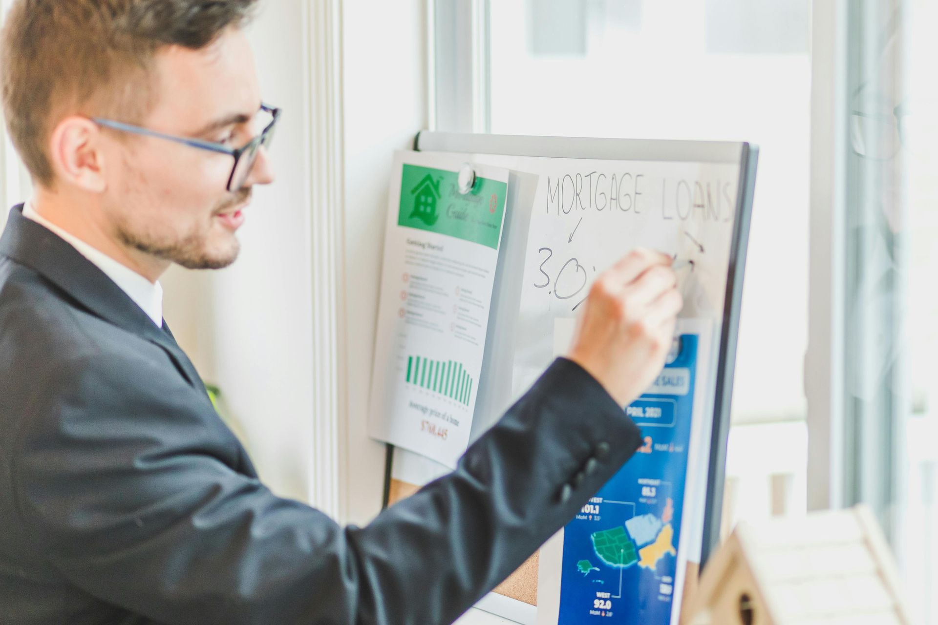 Man in glasses writing on a whiteboard beside a window in a bright office