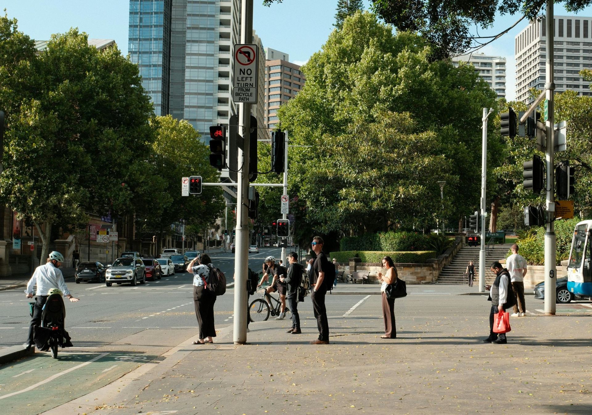 People waiting at a city crosswalk under traffic lights, with trees and tall buildings in the background.
