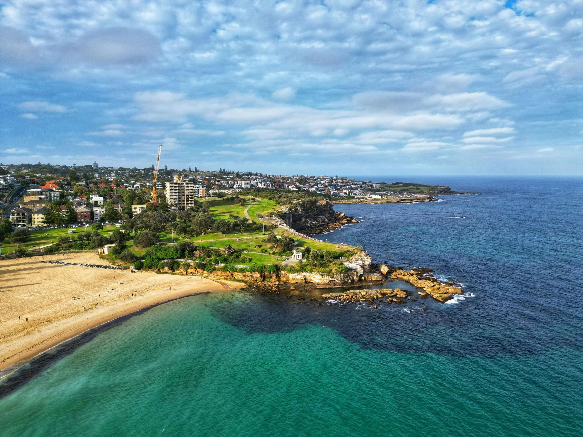 Coastal town with sandy beach, rocky shoreline, turquoise water, and blue sea under a cloudy sky