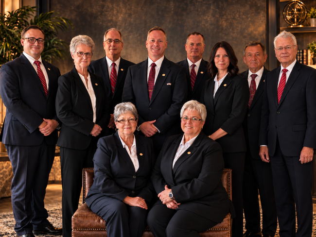 Group portrait of eight people in suits posing indoors against a dark backdrop