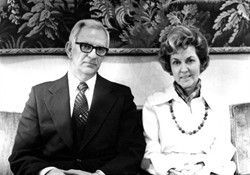 Two seated adults in formal attire, one in a suit and one in a white blouse with a necklace, in front of a mural