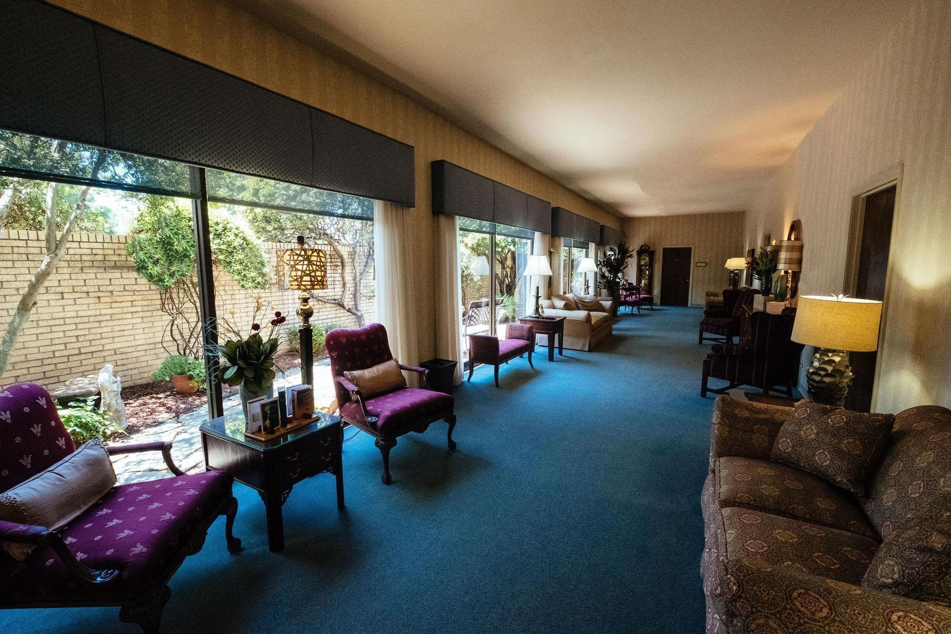 Long hallway lounge with blue carpet, large windows, purple chairs, and a table lamp