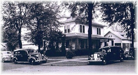 Vintage black-and-white street scene with parked cars in front of a large house and trees.