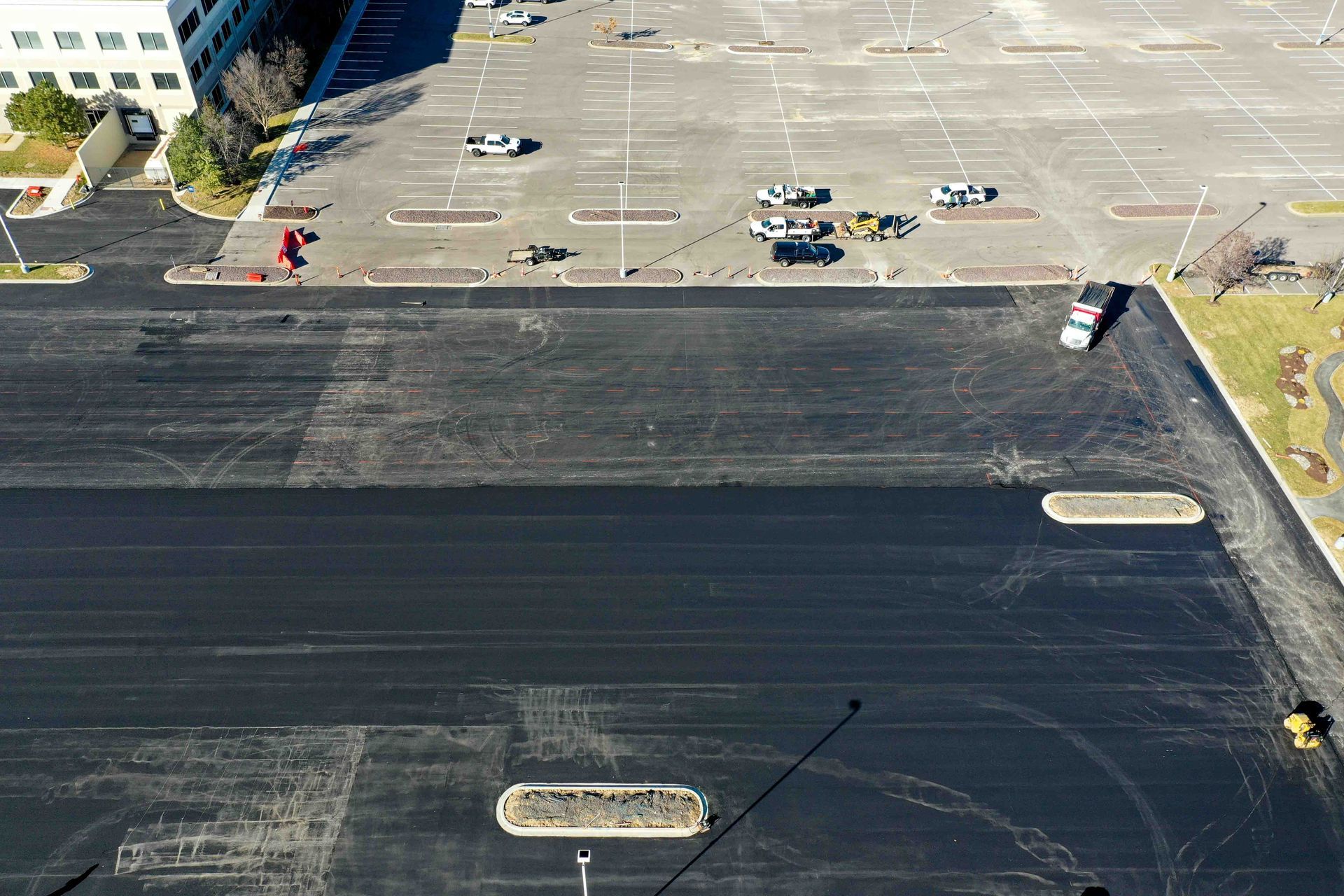 Aerial view of a large empty parking lot beside a dark paved surface with a few cars and people near a building.