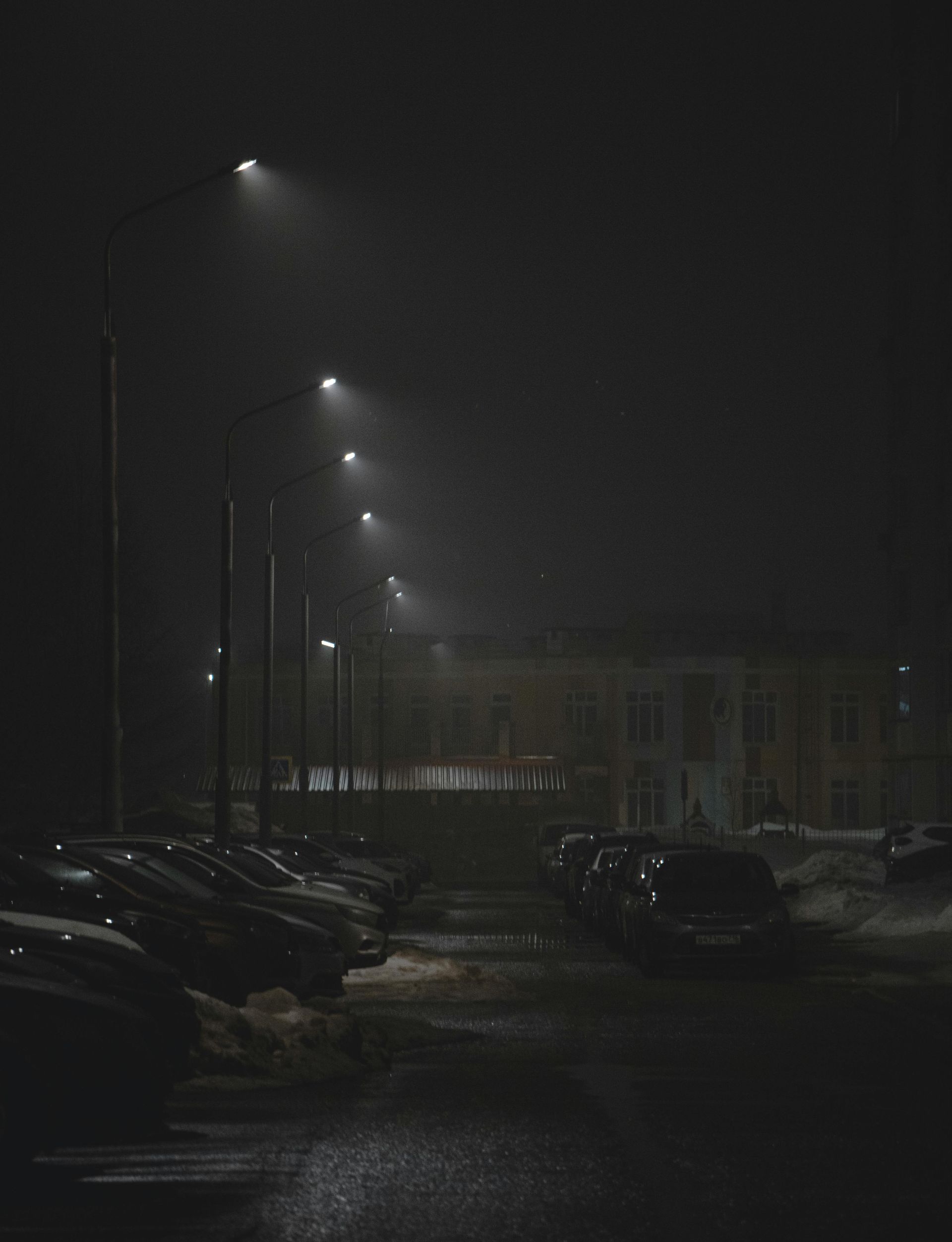 Night scene: row of streetlights illuminates a wet parking lot with parked cars, leading to a building in the fog.