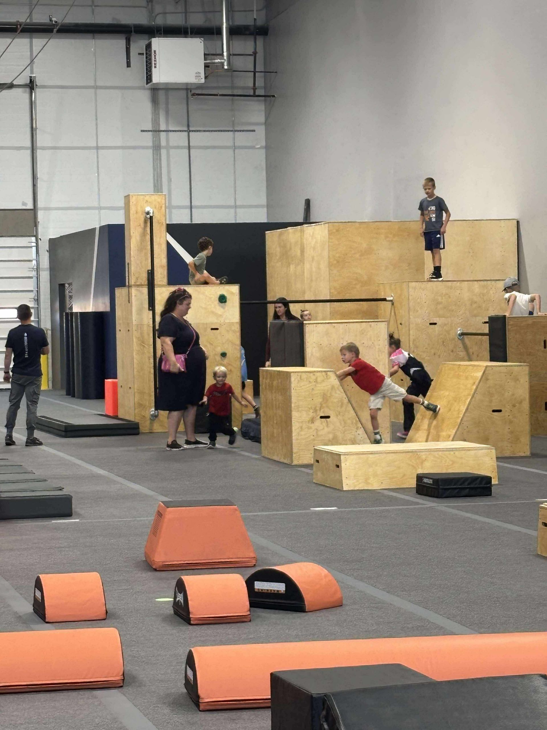 People in an indoor parkour gym, children climbing wooden structures, adults observing.
