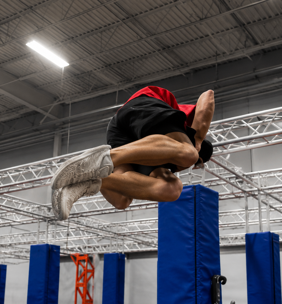 Man leaping over obstacles in an indoor training space, arms outstretched, leg extended. Black and white.