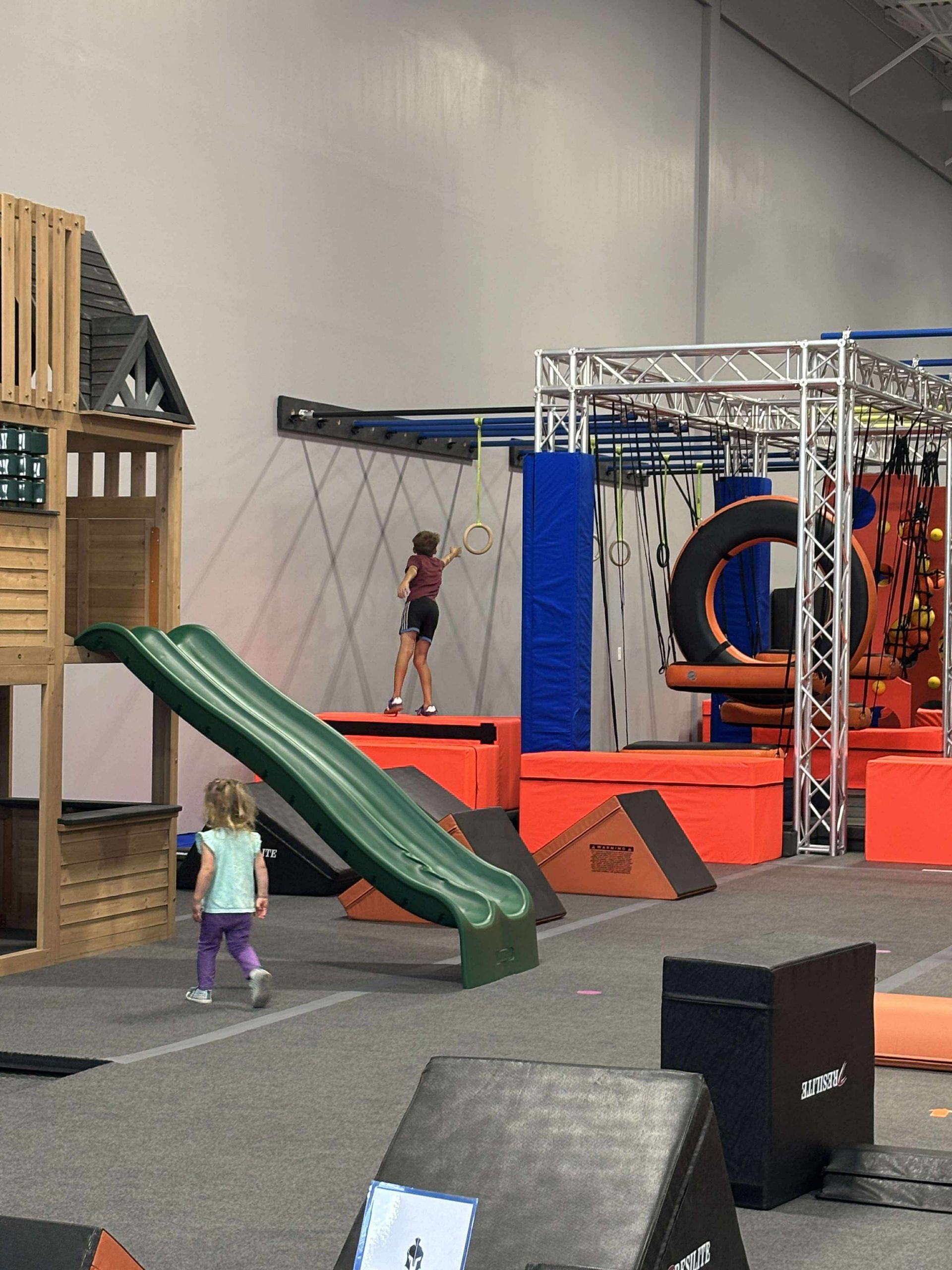 Children playing at indoor obstacle course with slide and climbing structures.