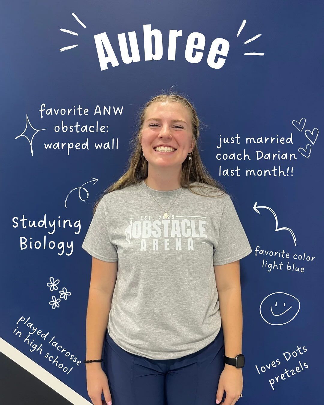 A woman smiles in front of a blue wall with facts about her. She's wearing a grey shirt that says