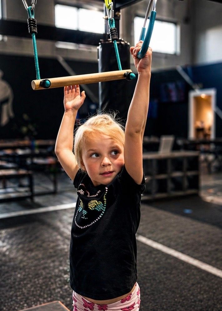 Child holding onto gym rings in a fitness gym, arms raised, wearing a black shirt and pink shorts.