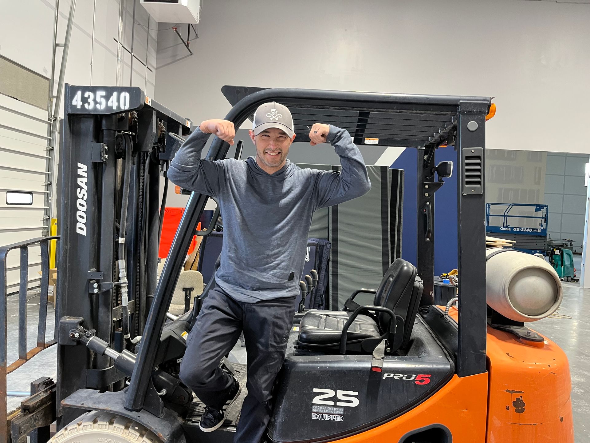 Man flexing arms, standing in orange forklift cab. Gray shirt, dark pants, and baseball cap, indoors.