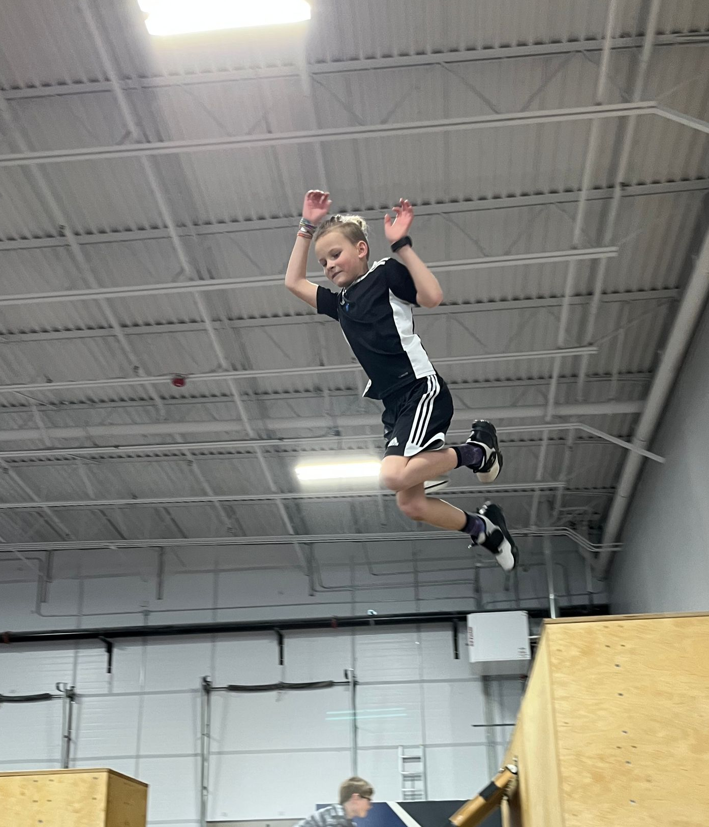 Boy balancing on a beam in a gym; instructor watches. Blue climbing wall in background.