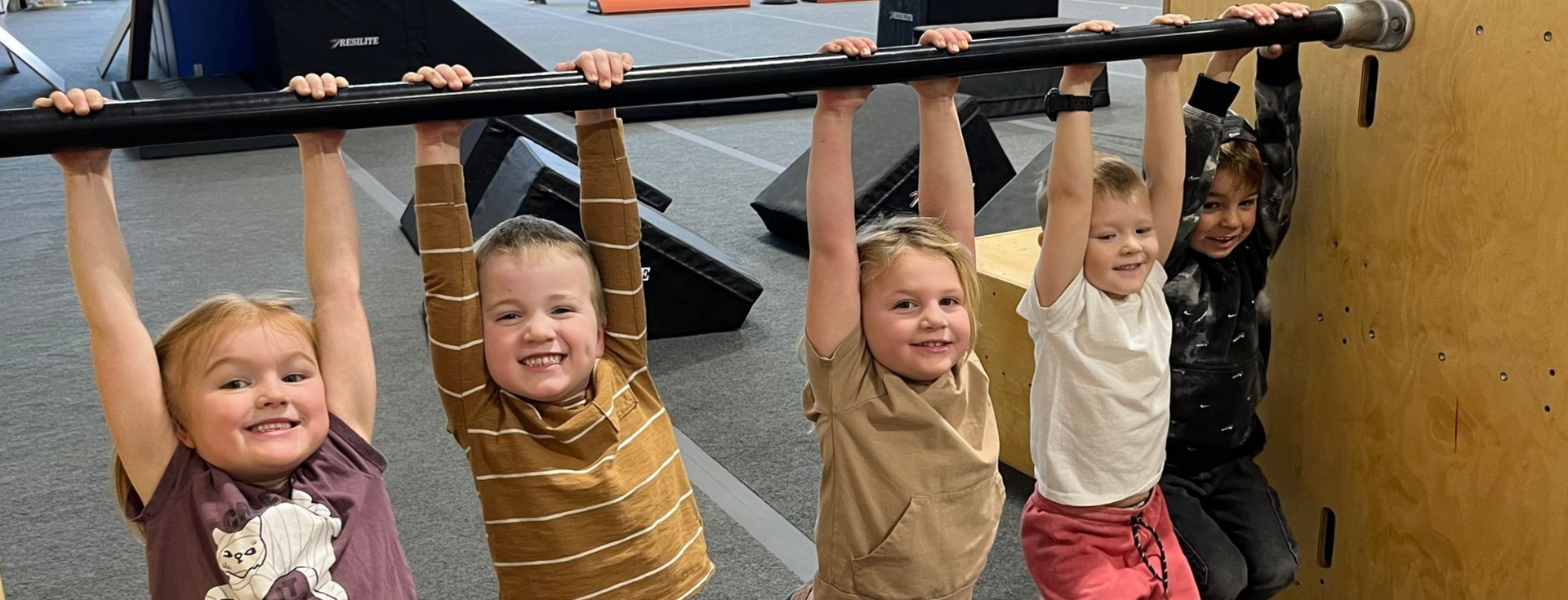 Children hanging from a horizontal bar in a gym, smiling and playing together.