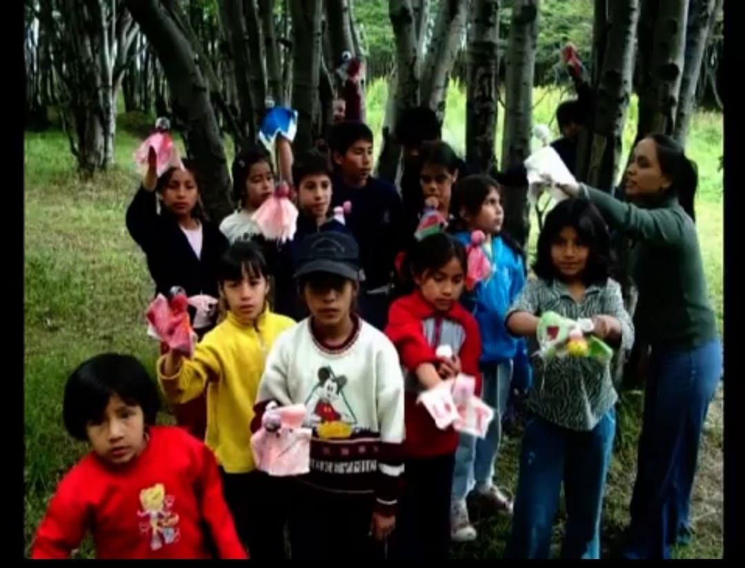 Niños y una mujer en un bosque, sosteniendo pequeños paquetes de color rosa y blanco.